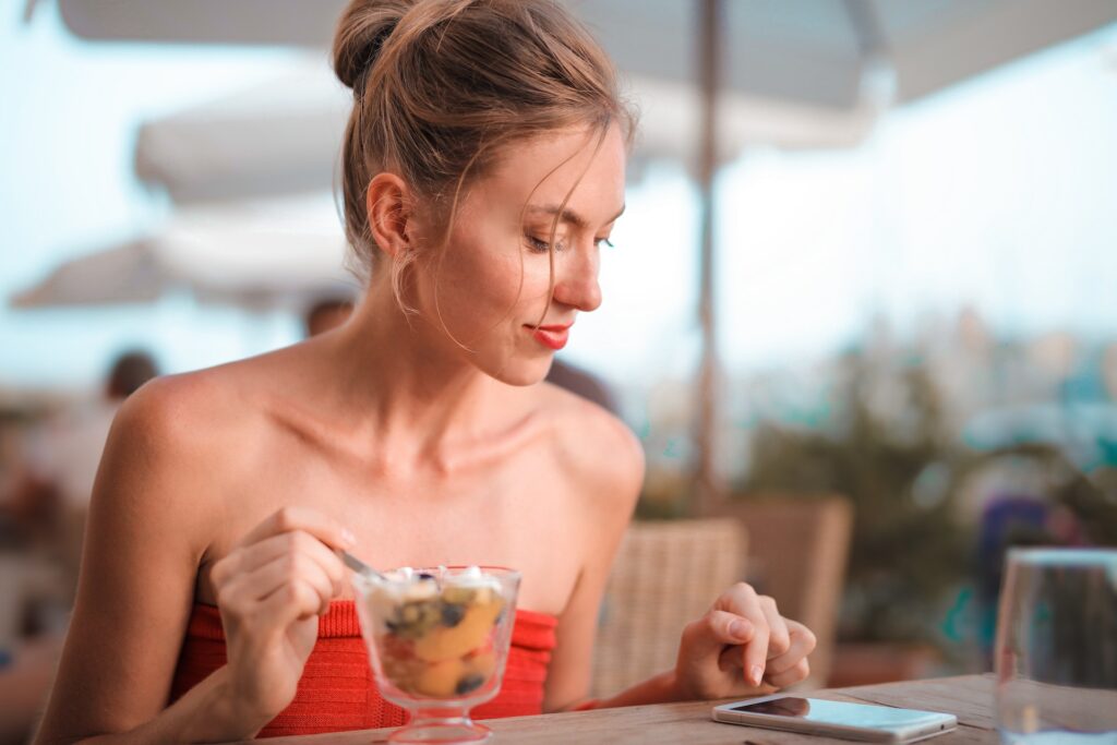 Young lady eating a dessert at an outdoor restaurant while also playing with her mobile phone, which sits on the table