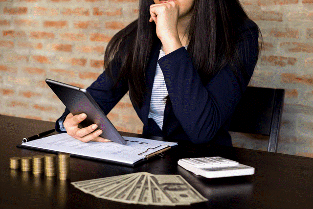 A woman holding a tablet sits at a table with a calculator, dollars, and a clipboard; she's a restaurant owner who's decided to monitor her cost of good solds