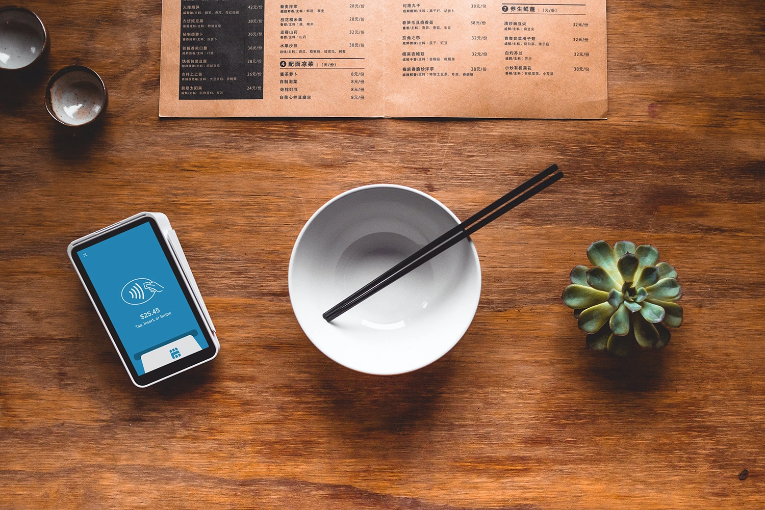 A phone, bowl, and chopsticks on a wooden table amidst menu pricing considerations.